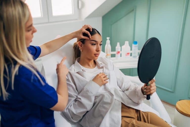A woman sitting, looking into a round mirror in her hand, talking to a doctor about her eyebrows